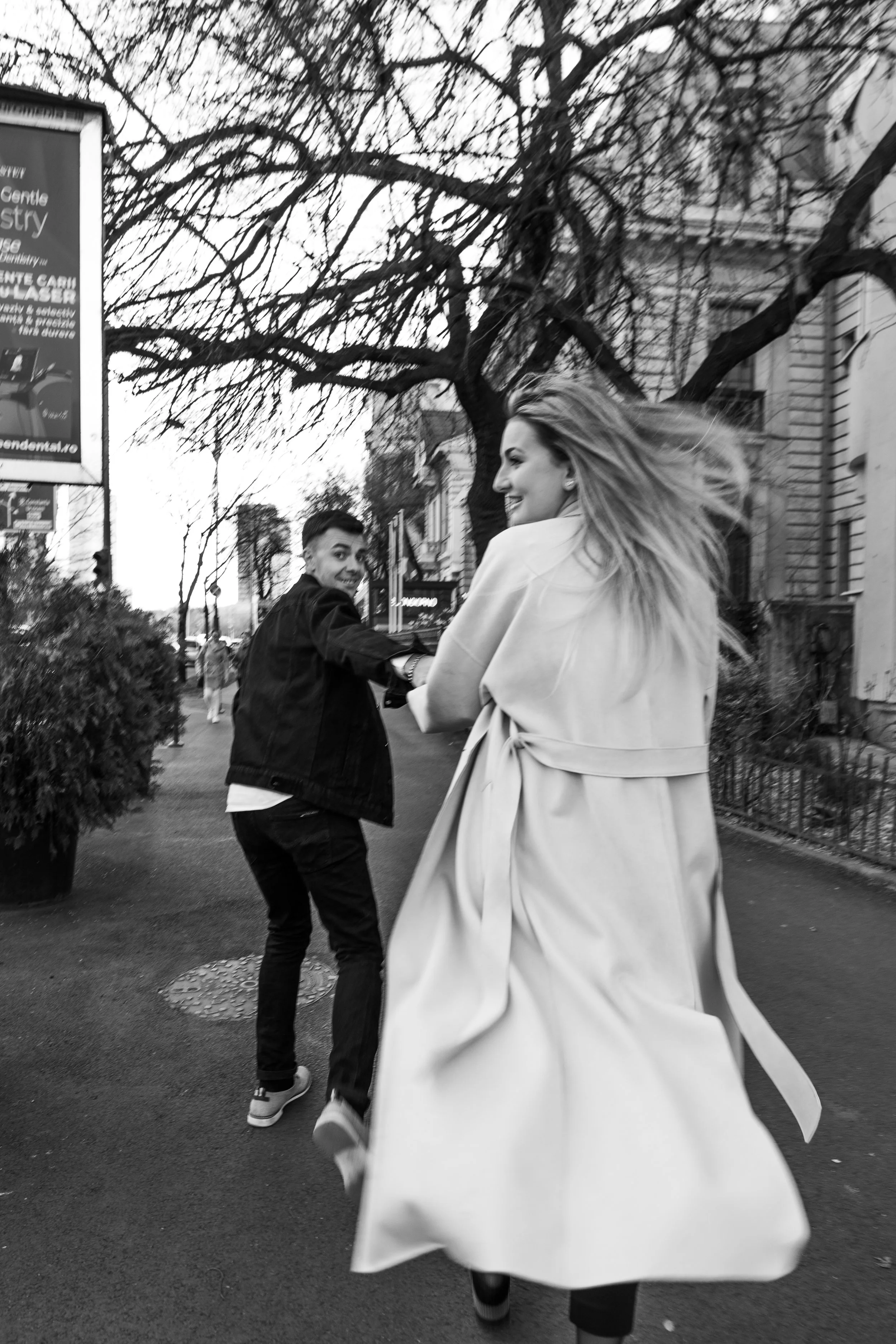A black-and-white photo of a woman and a man holding hands and smiling as they walk down a city street, with leafless trees and buildings in the background.