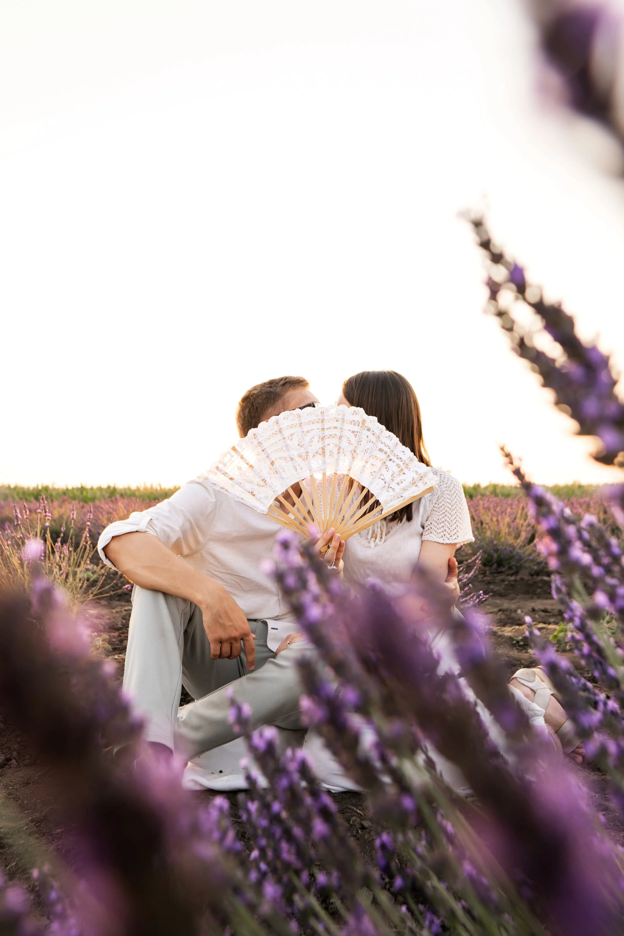 A couple sitting in a lavender field at sunset, with the man holding a lace fan and both sharing a kiss