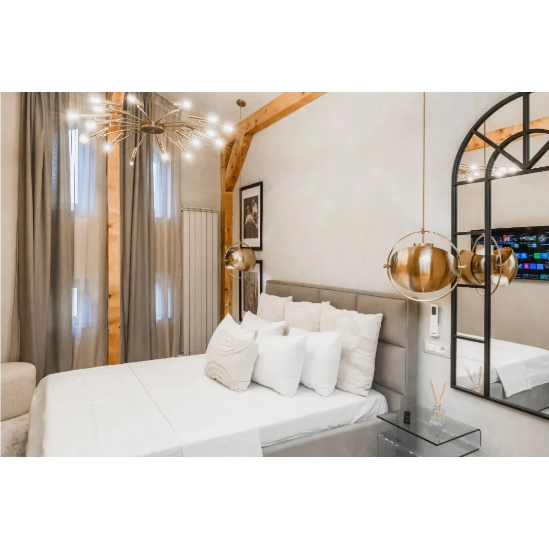 Modern bedroom with a large bed, multiple pillows, wooden beams, stylish hanging lamps, a framed picture, a black mirror, and a flat-screen TV on the wall.