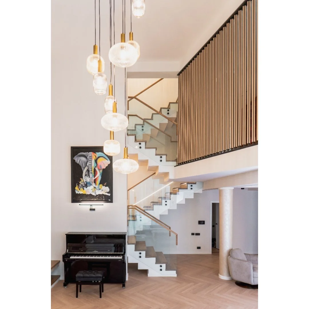 Interior of a modern home with a staircase, hanging pendant lights, a piano with a stool, a colorful painting on the wall, and a curved armchair at the corner.