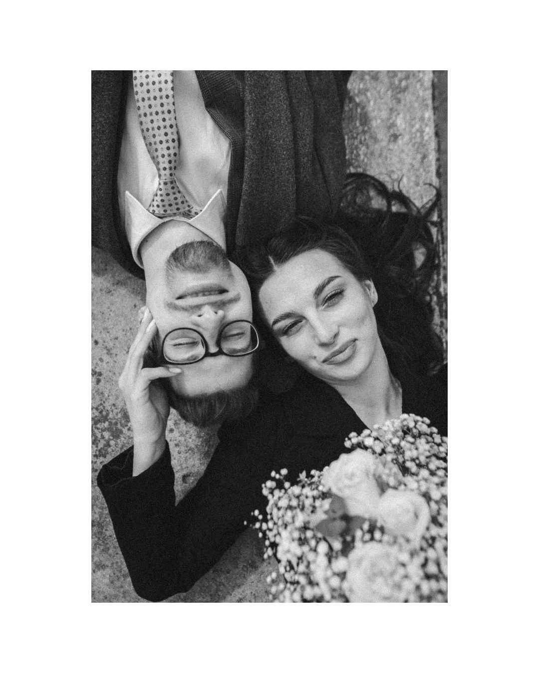 A black-and-white photo of a man and woman lying on the ground, taking a selfie together. The man is smiling with glasses, and the woman is sticking out her tongue with her eyes slightly squinted. There are flowers near the woman, and the background shows both their clothing and the ground they are lying on.