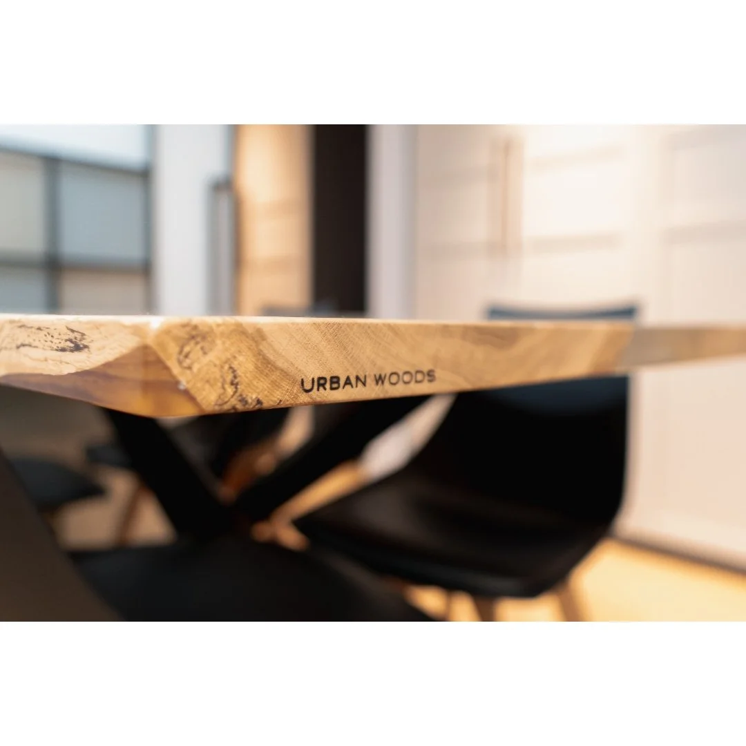 Close-up of a wooden table with the words "URBAN WOODS" printed on the edge, with black chairs and a bright, modern interior in the background.