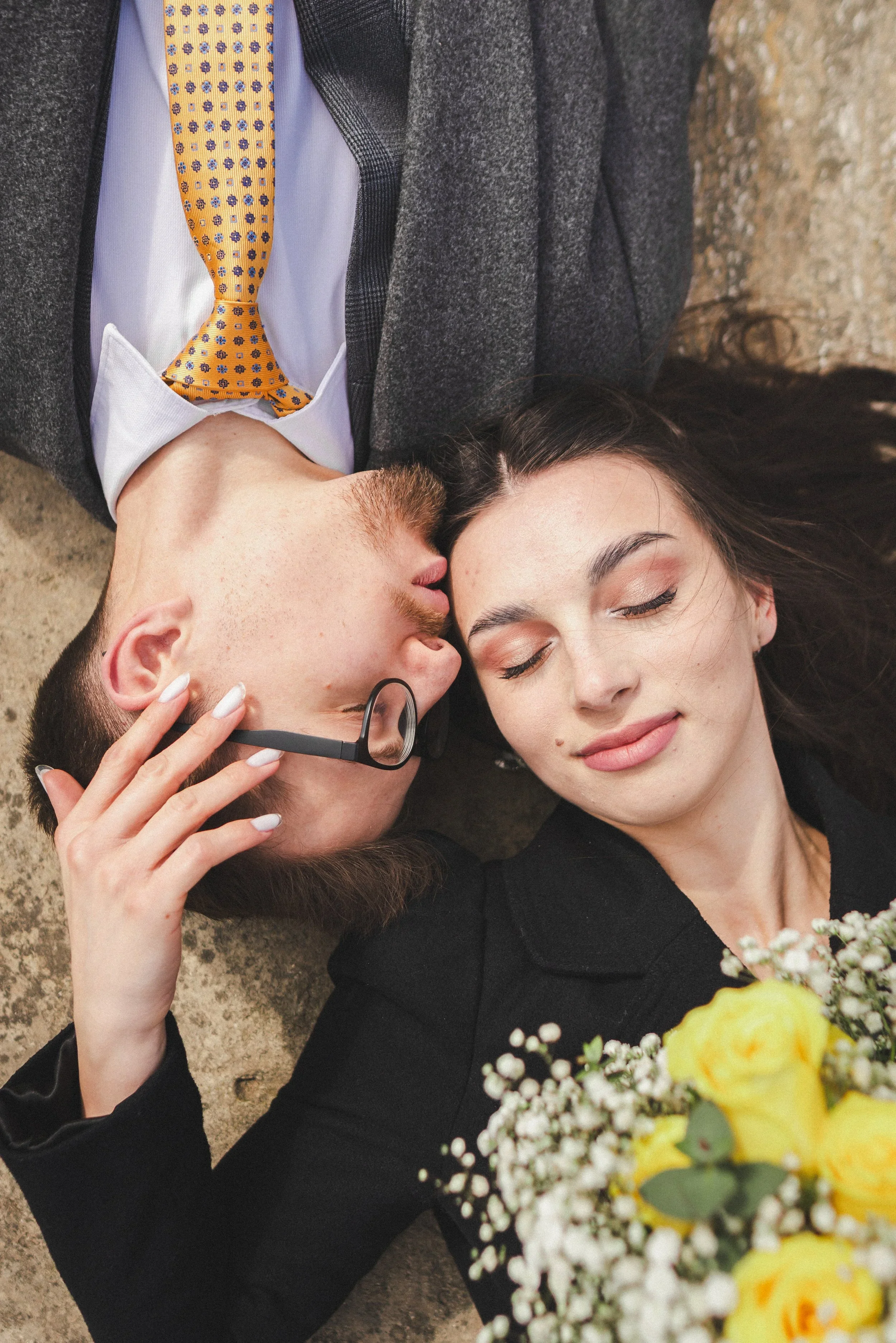 A couple lying on the floor, with the man wearing glasses and a suit, and the woman holding a bouquet of yellow and white flowers. They are close together, with their heads touching and eyes closed.