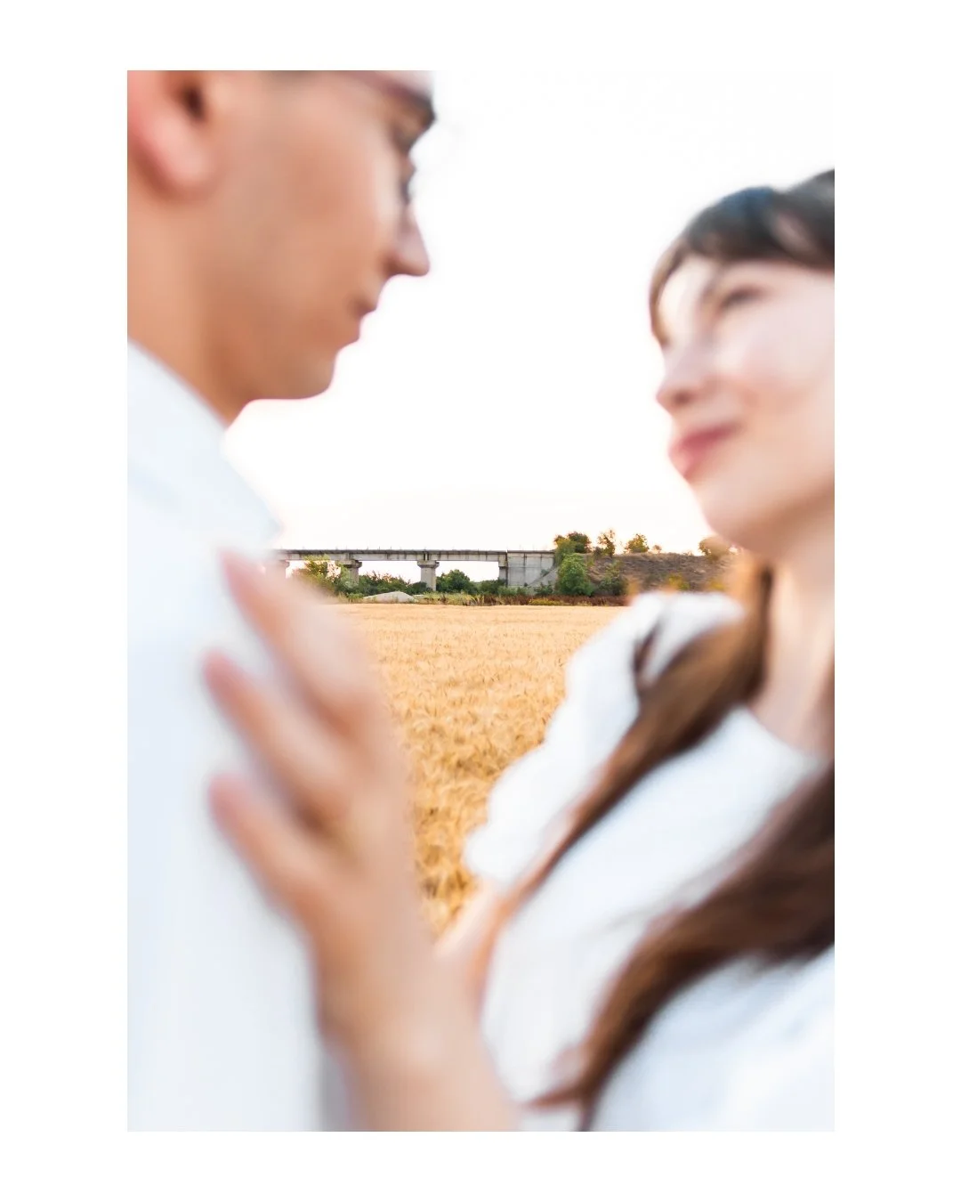 Two people standing in a field with a train bridge in the background, close together and slightly out of focus.