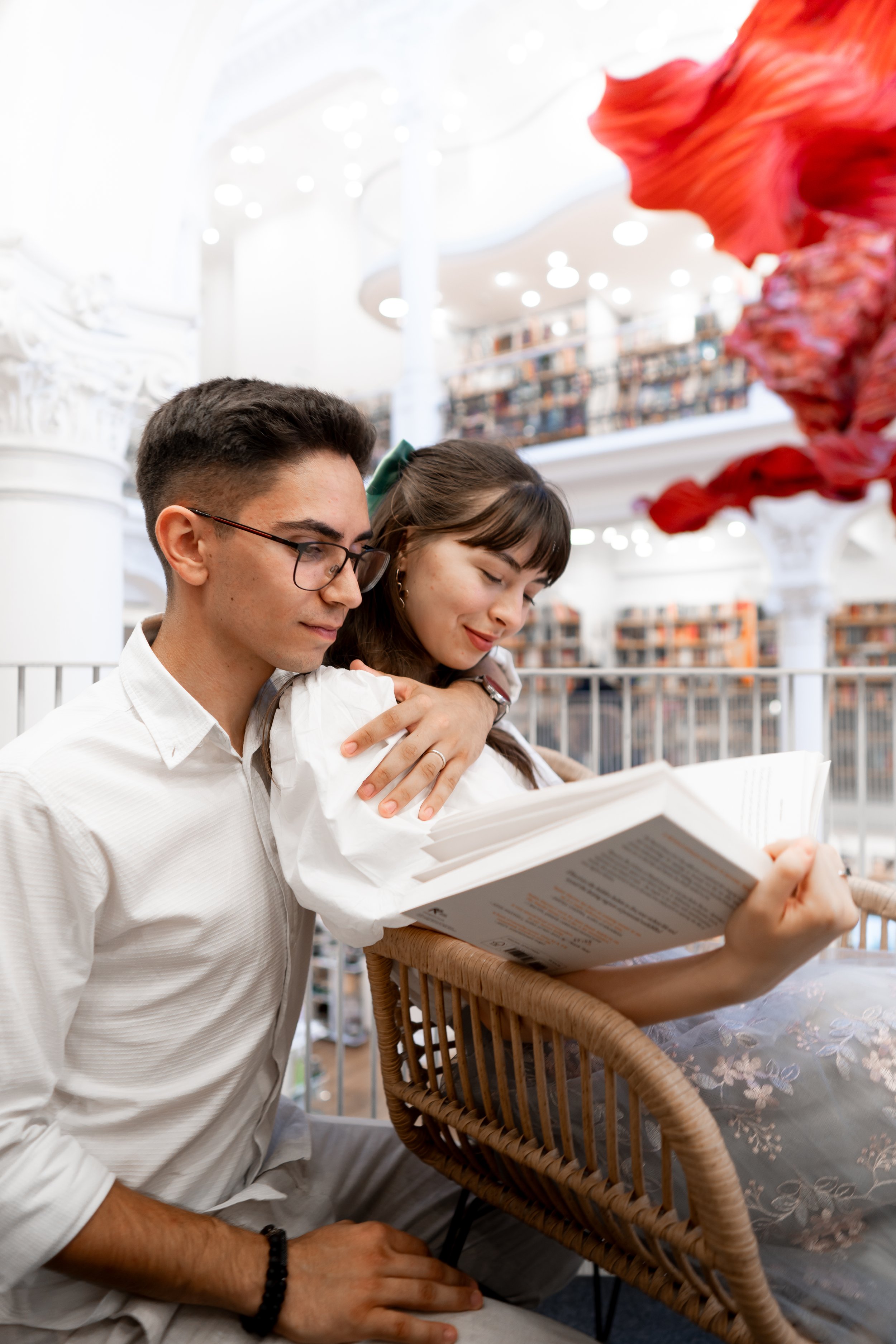 A young man and woman sitting in a library, reading a book together. The woman is leaning on the man's shoulder, and both are smiling softly. The background shows bookshelves and a bright, modern interior.