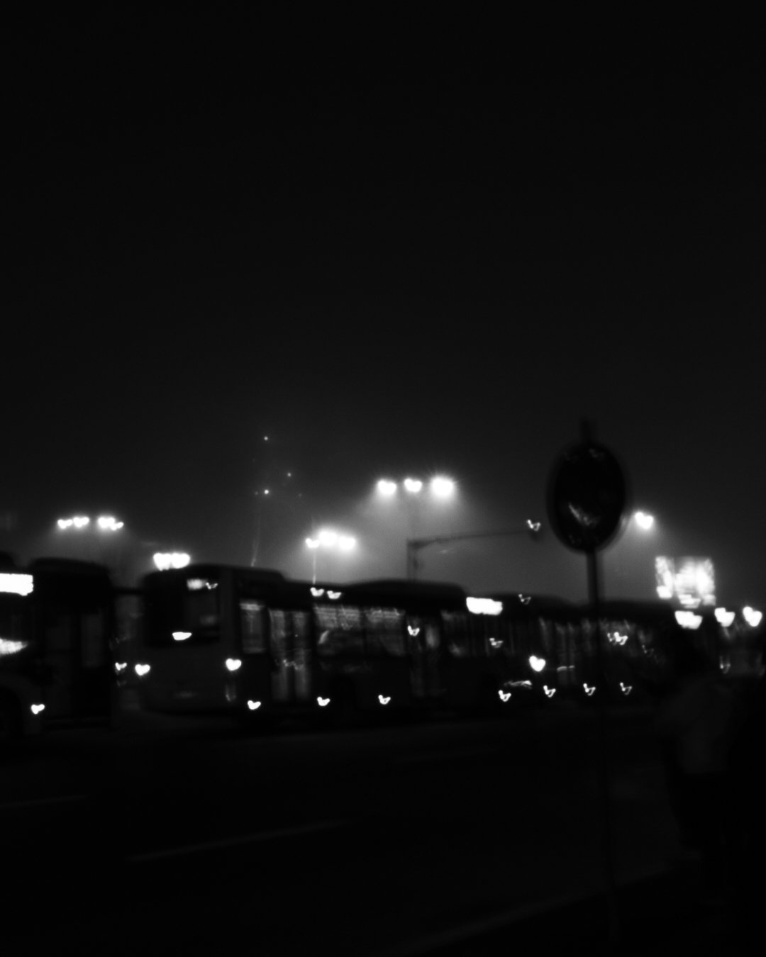 Nighttime scene of a bus terminal with multiple buses parked in rows, illuminated by overhead lights, with a hazy sky and a sign in the foreground.