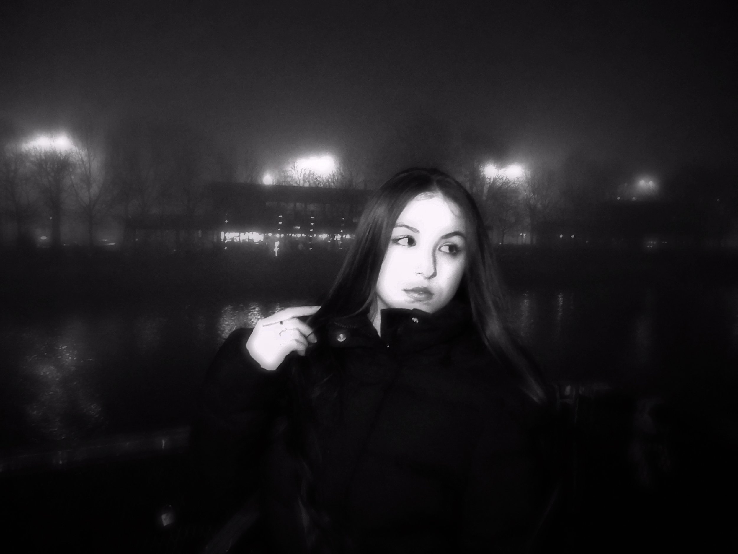 Black and white photo of a young woman with long hair, wearing a dark coat, standing outdoors at night near water with faint city lights and trees in the background.