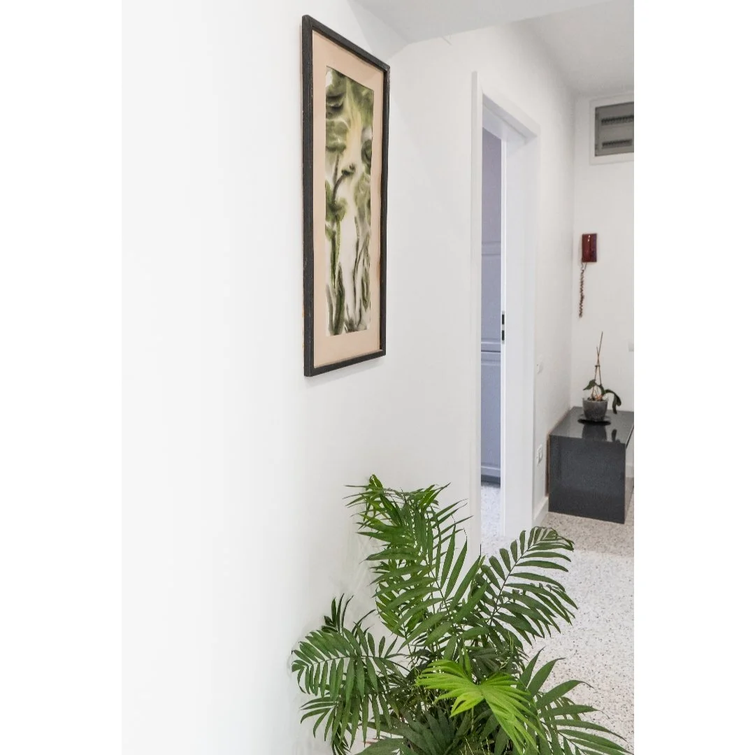 Interior scene with a white wall, a framed abstract art piece, a green potted plant, and a small black table with a pot and plant on it.