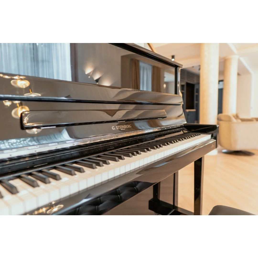Black G. Steinberg piano in a well-lit living room with curtains and beige sofa in the background.