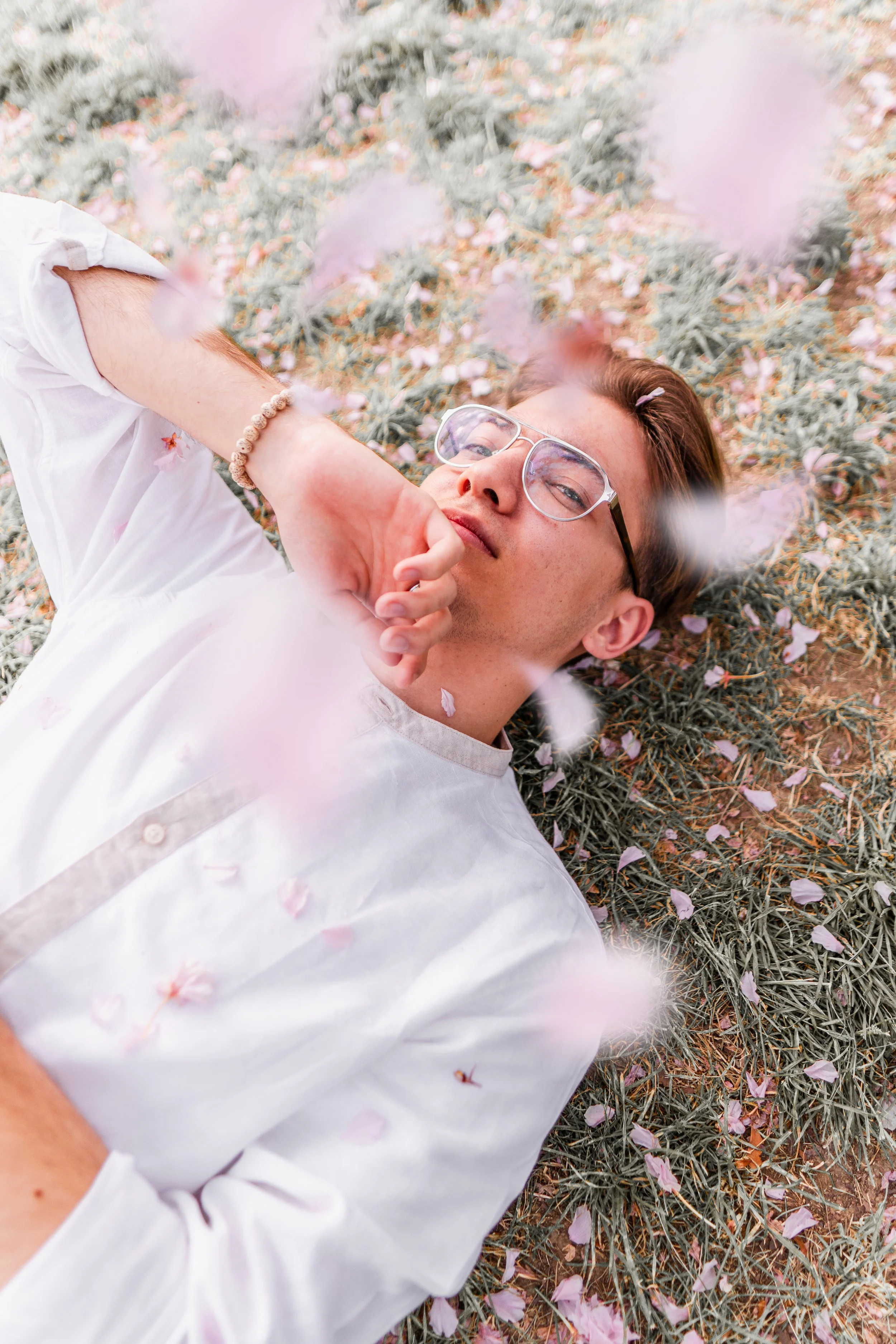 A young man with glasses lying on grass with pink flower petals scattered around, surrounded by falling petals.