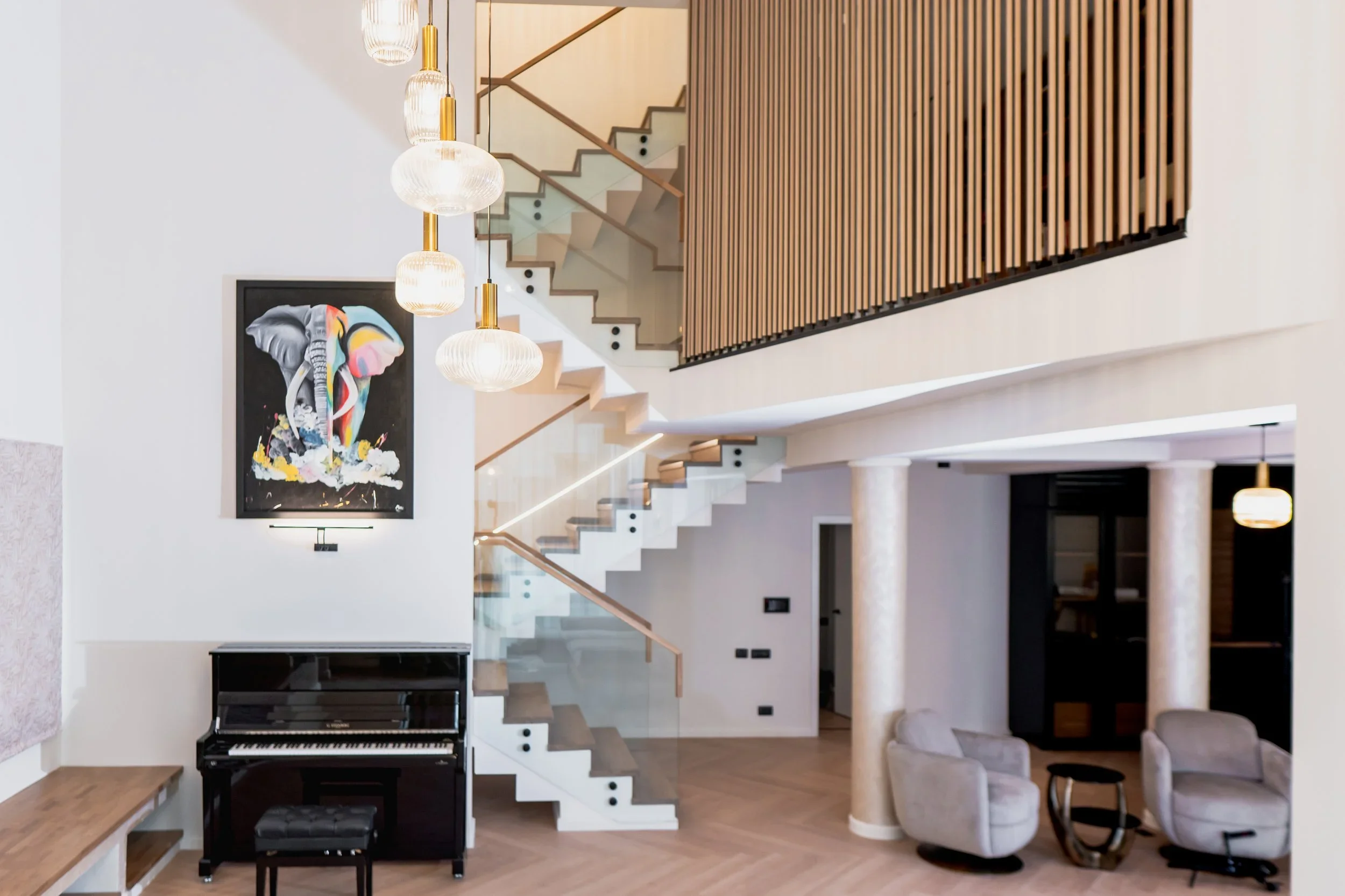 Modern living room with white walls, wooden flooring, two beige armchairs, a small black side table, a black upright piano with a matching stool, a colorful elephant painting on the wall, a staircase with glass railing, wooden accents, and pendant lighting.