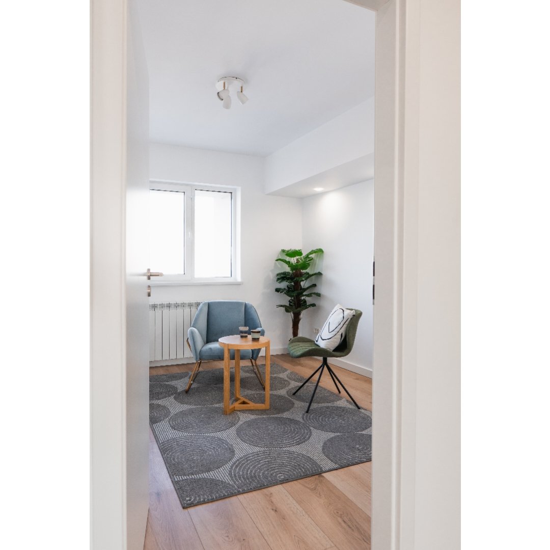 A cozy sitting area with two chairs, a small wooden table with cups, a patterned rug, a potted plant, and a window in a white-walled room.