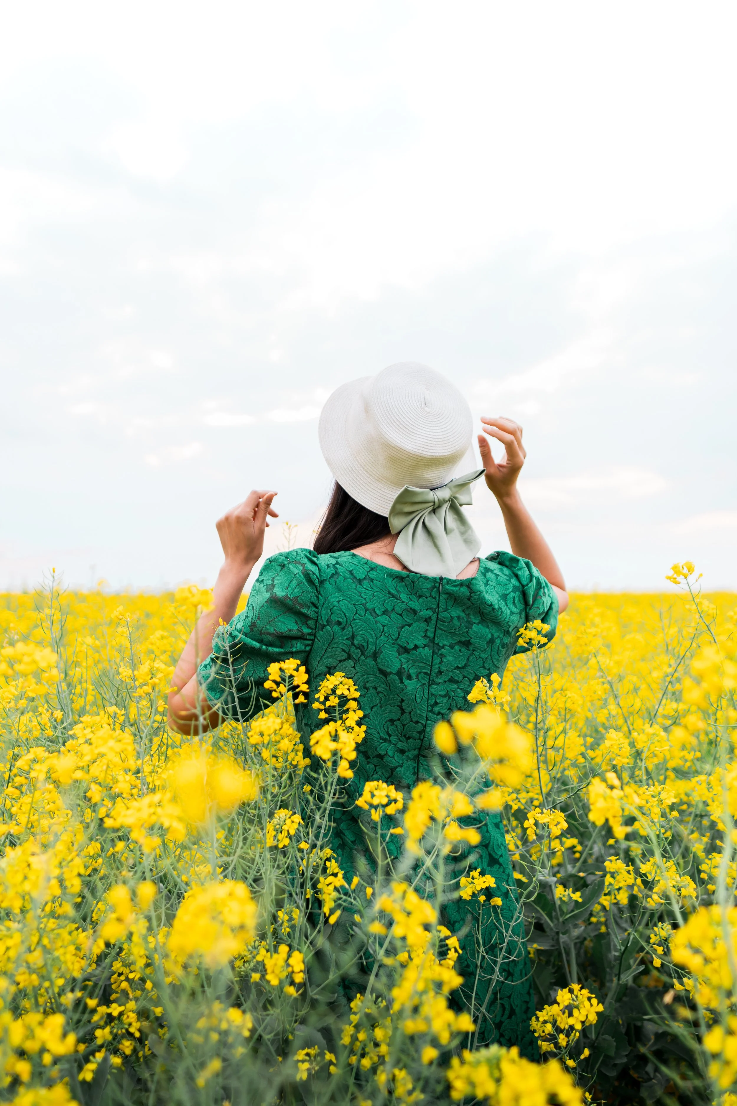 A woman in a green dress and white sun hat with a green bow walking in a yellow flower field under a cloudy sky.