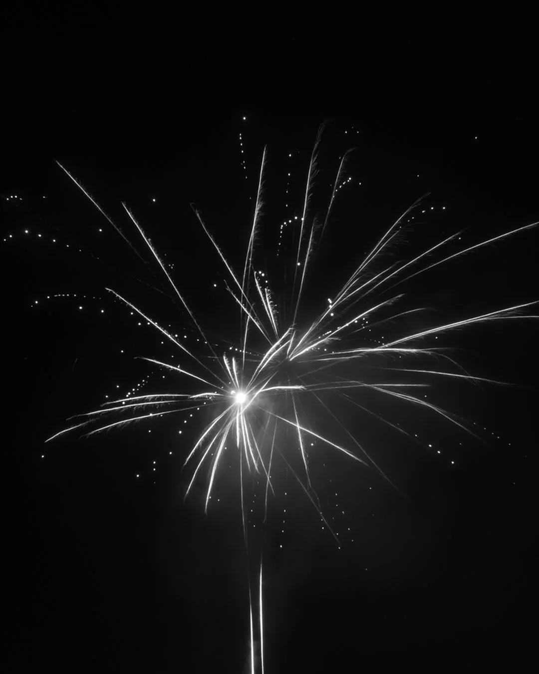 Black and white photograph of a firework explosion in the night sky with streaks and dots of light radiating from the center.