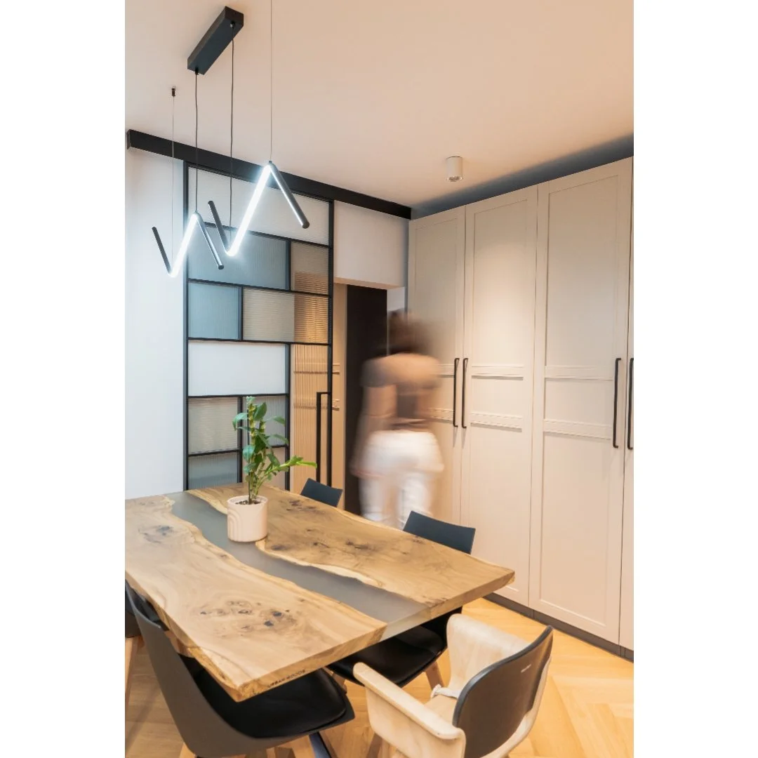 Modern dining room with a wooden table, black chairs, a potted plant, geometric black and glass room divider, white built-in cabinetry, and a ceiling light fixture with a zigzag design.