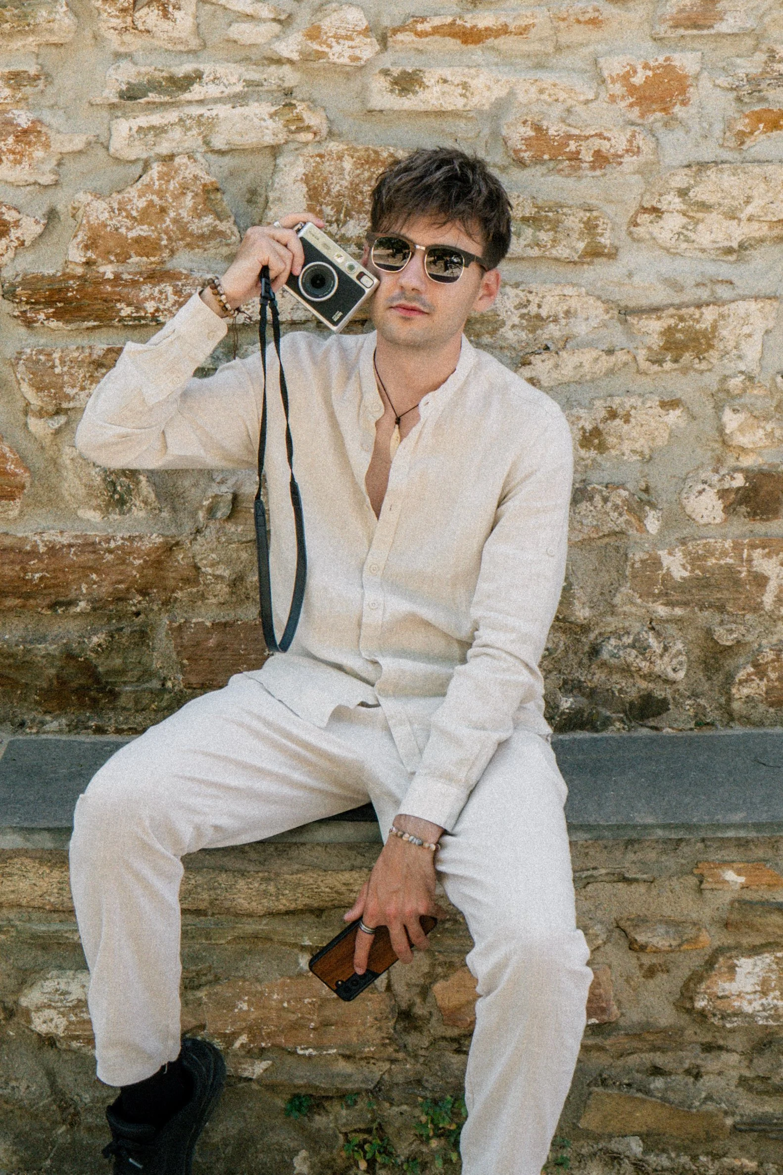 A young man with dark hair, wearing sunglasses and a beige outfit, is sitting against a rustic brick wall. He is holding a vintage camera in one hand and a smartphone in the other, with a relaxed and stylish pose.