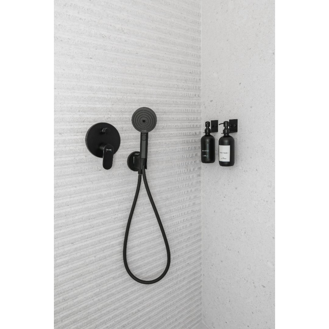 Black shower fixtures, including a handheld showerhead, wall-mounted handle, and two wall-mounted bottles, are installed on a textured wall in a modern bathroom.