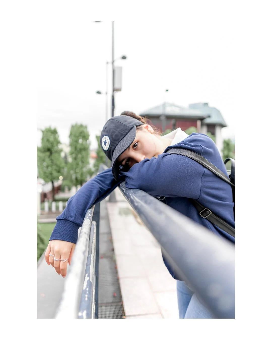 Young woman leaning on a black metal railing outdoors, wearing a blue hoodie, a black Converse cap, and carrying a backpack, looking into the camera with a contemplative expression. Trees and a building are visible in the background.