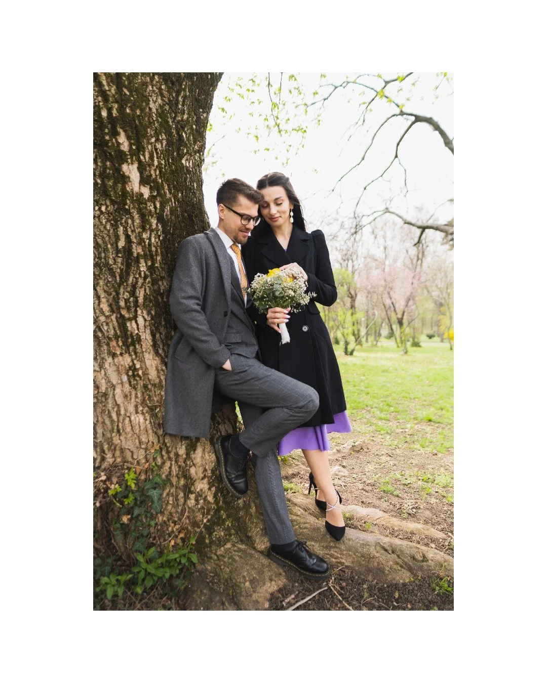 A man and woman dressed elegantly, leaning against a large tree in a park, with the woman holding a bouquet of flowers and both looking down, surrounded by budding trees and greenery.