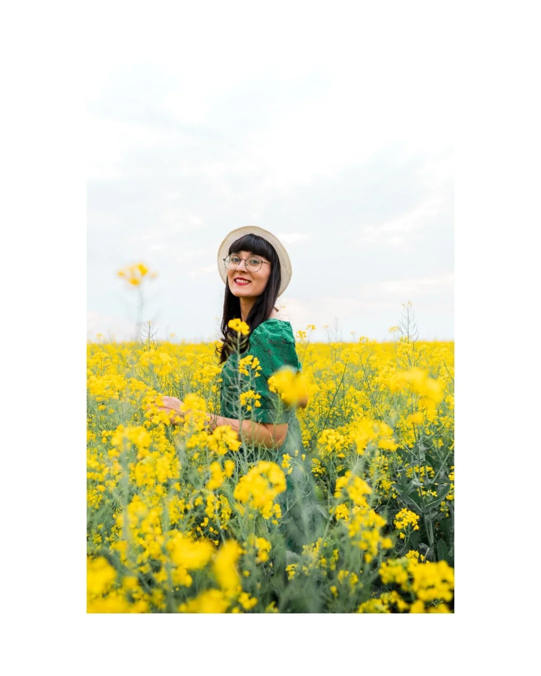 A woman with glasses and a hat standing in a field of yellow flowers, smiling, with a cloudy sky in the background.