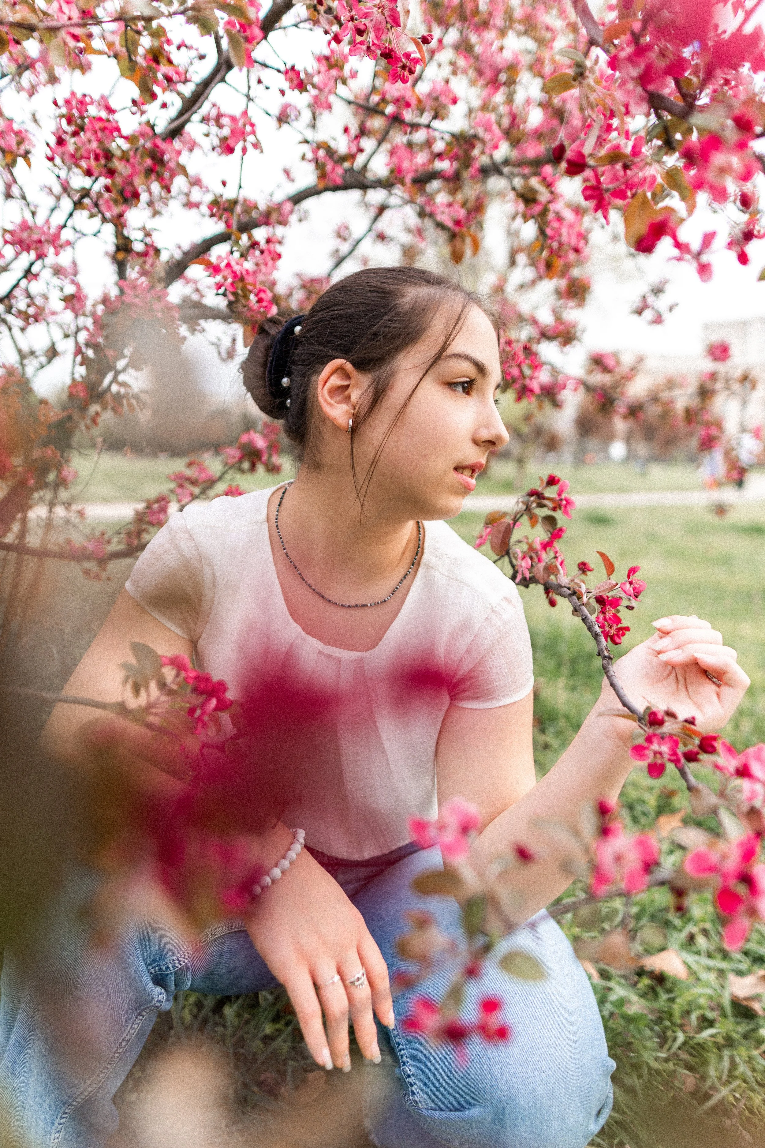 A young woman with dark hair in a bun is sitting outdoors among pink blooming branches, wearing a light top and jeans, looking thoughtfully at the flowers.