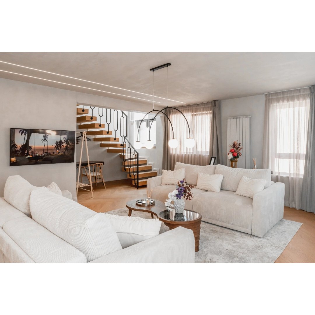 A modern living room with white sofas, a wooden coffee table with a vase of flowers, large windows with sheer curtains, a staircase with wooden steps and black railing, a wall-mounted TV displaying a sunset scene with palm trees, and decorative lighting fixtures.