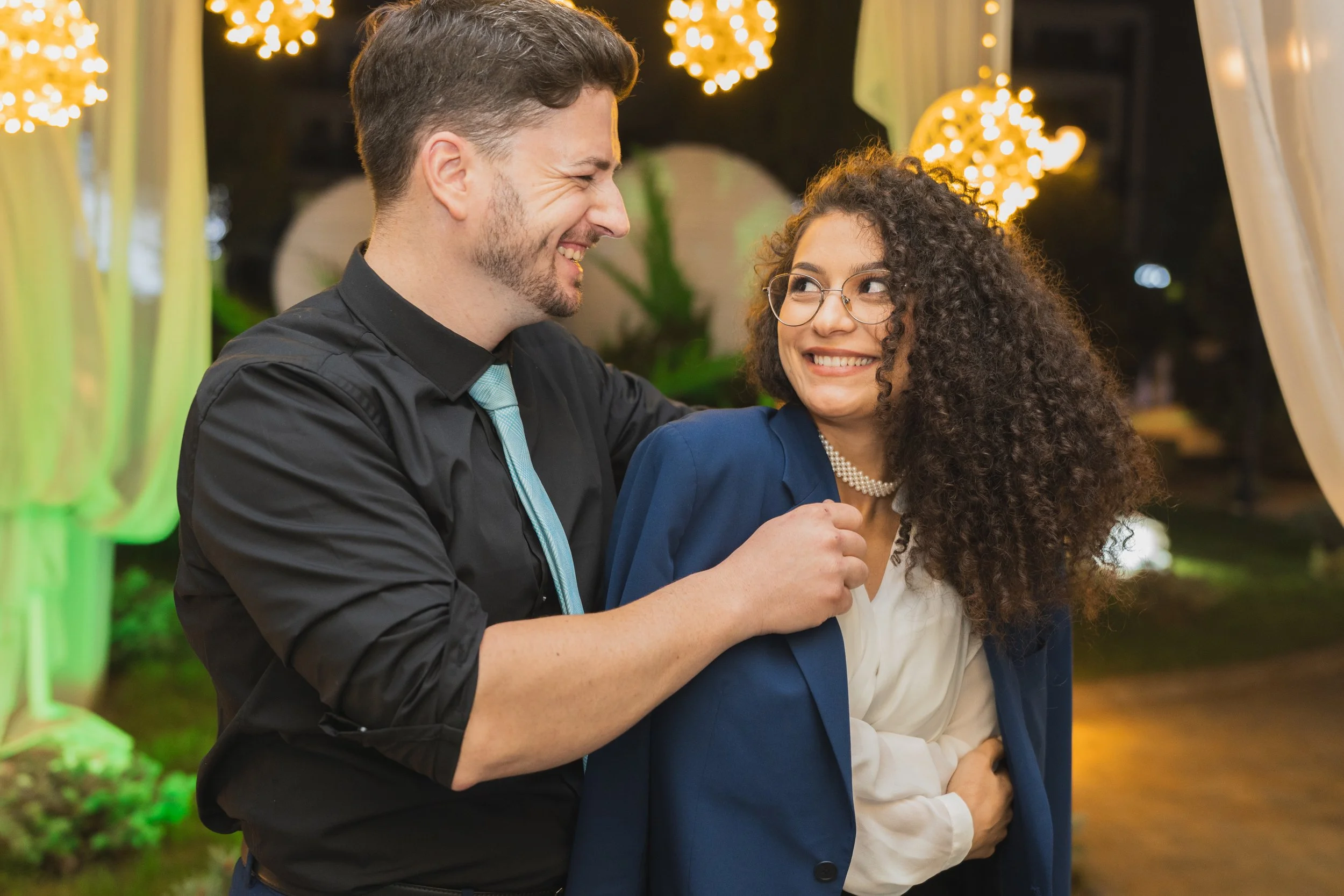 A man and woman smiling at each other at a festive indoor event, with decorative lights in the background.