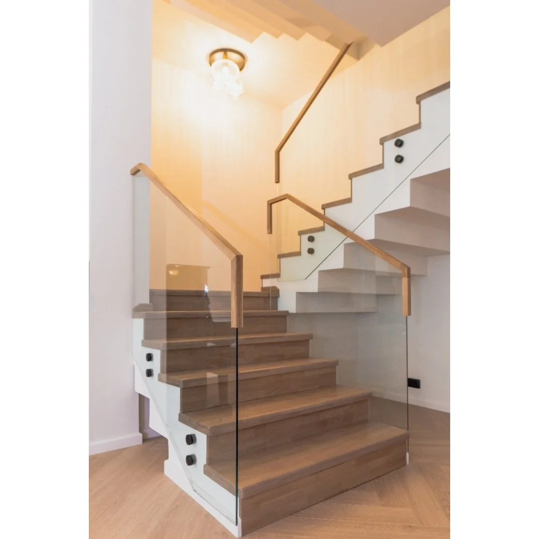 Modern staircase with wooden steps and glass balustrade, leading to an upper floor, in a well-lit interior with light-colored walls and ceiling.