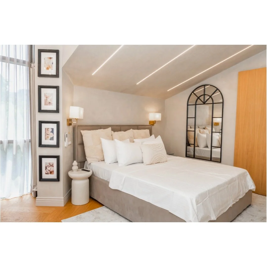 A modern bedroom with a bed, multiple pillows, a nightstand, wall-mounted lamps, a large mirror, and framed artwork, featuring neutral tones and natural light.