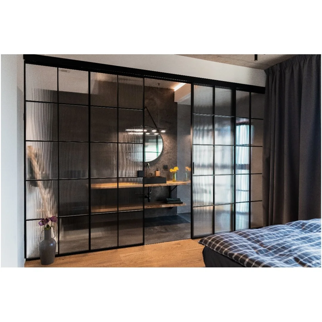 Modern bedroom with a glass sliding door separating the sleeping area from the bathroom. The room has a bed with checkered bedding, a potted purple flower on a stool, and a vanity with a round mirror, candles, and flowers.