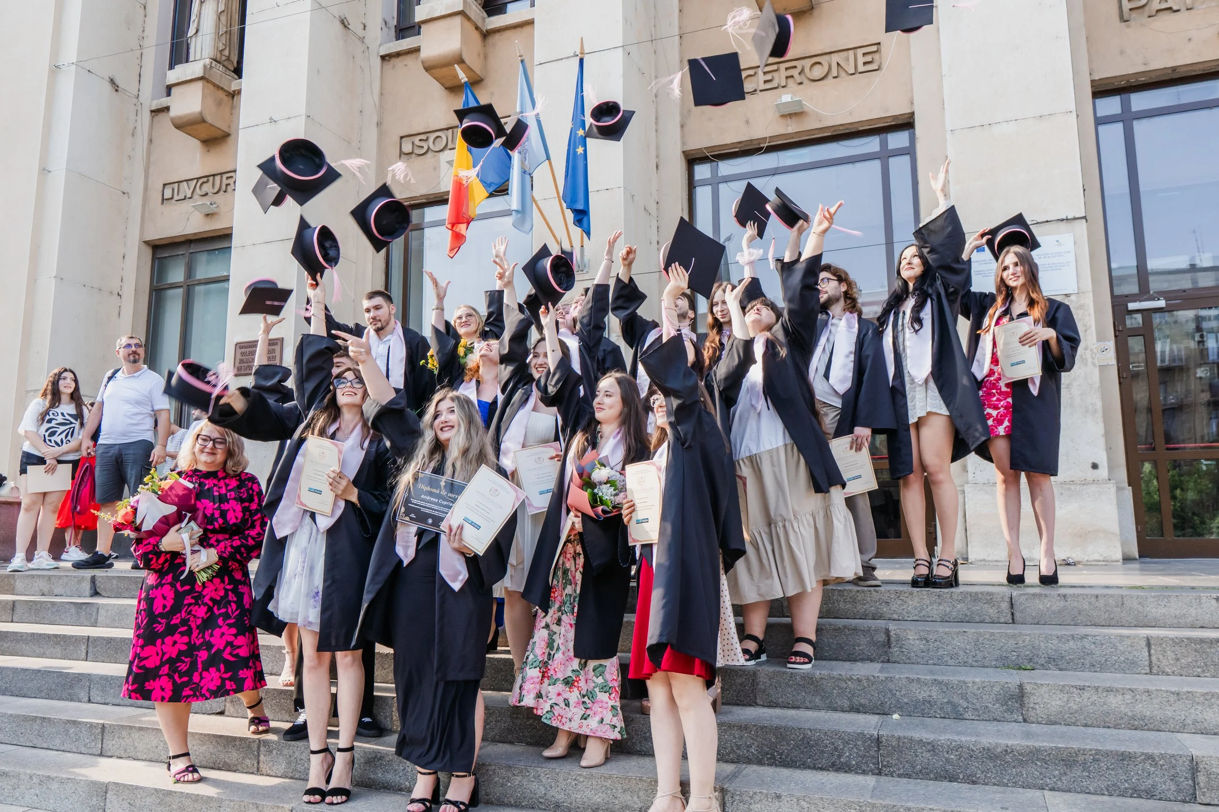 Group of graduates in caps and gowns celebrating on steps outside a building with flags