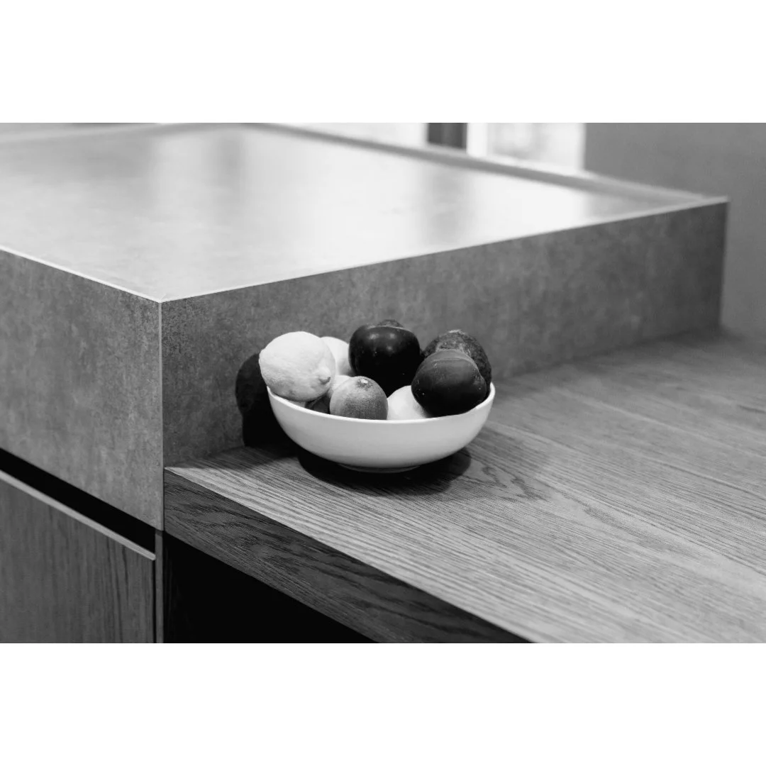 A white bowl filled with assorted fruits on a wooden table near a minimalist counter.
