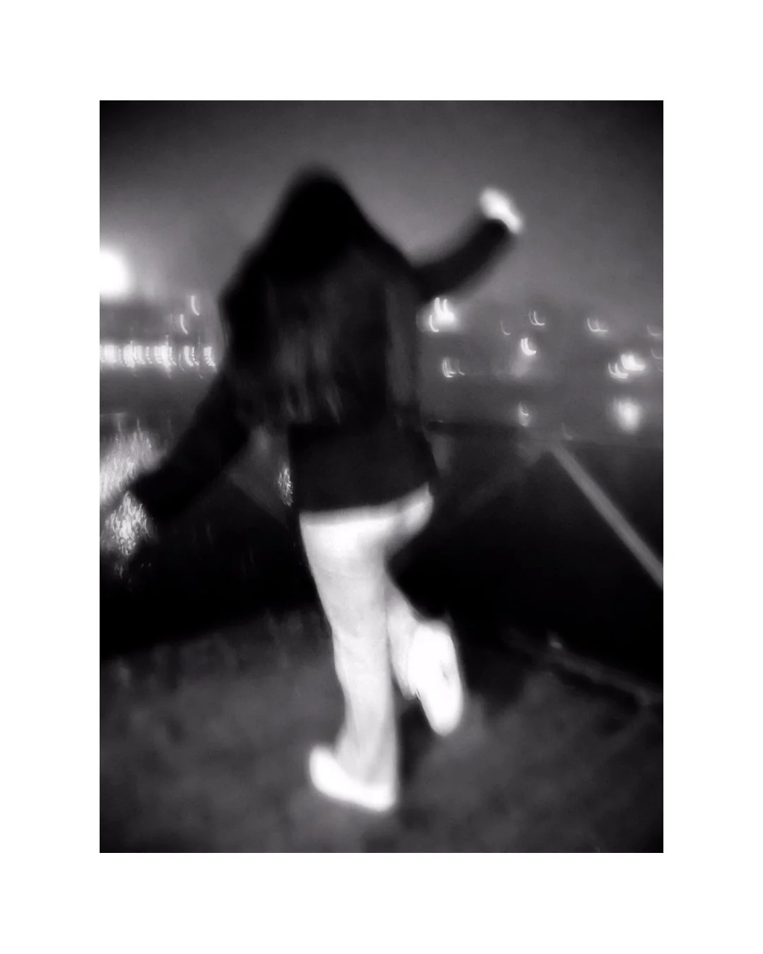 Person standing on a bridge at night, facing away, taking a selfie with a smartphone, with city lights and water below.