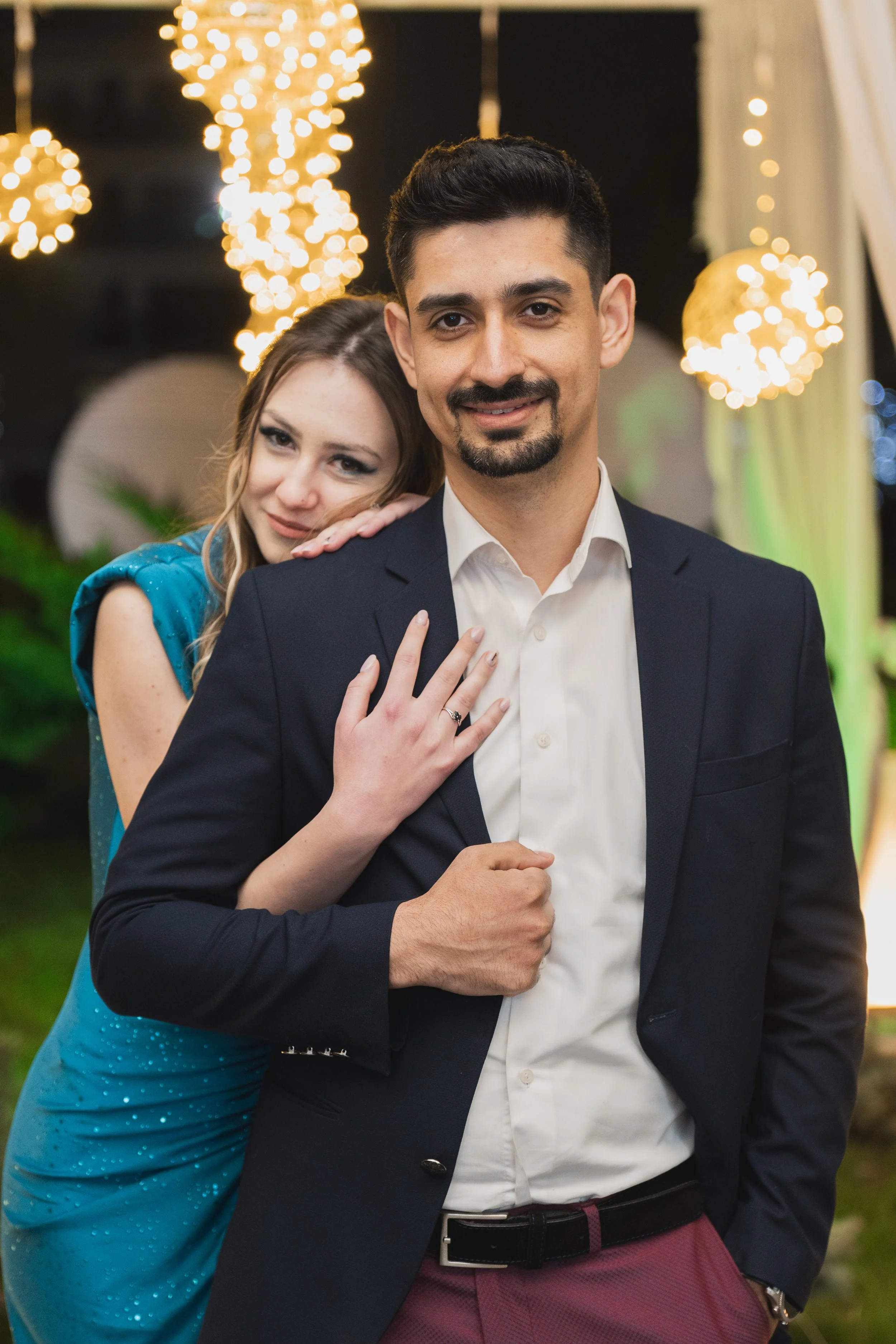 A man in a suit and a woman in a blue dress smiling at the camera at an evening event with blurred string lights in the background.