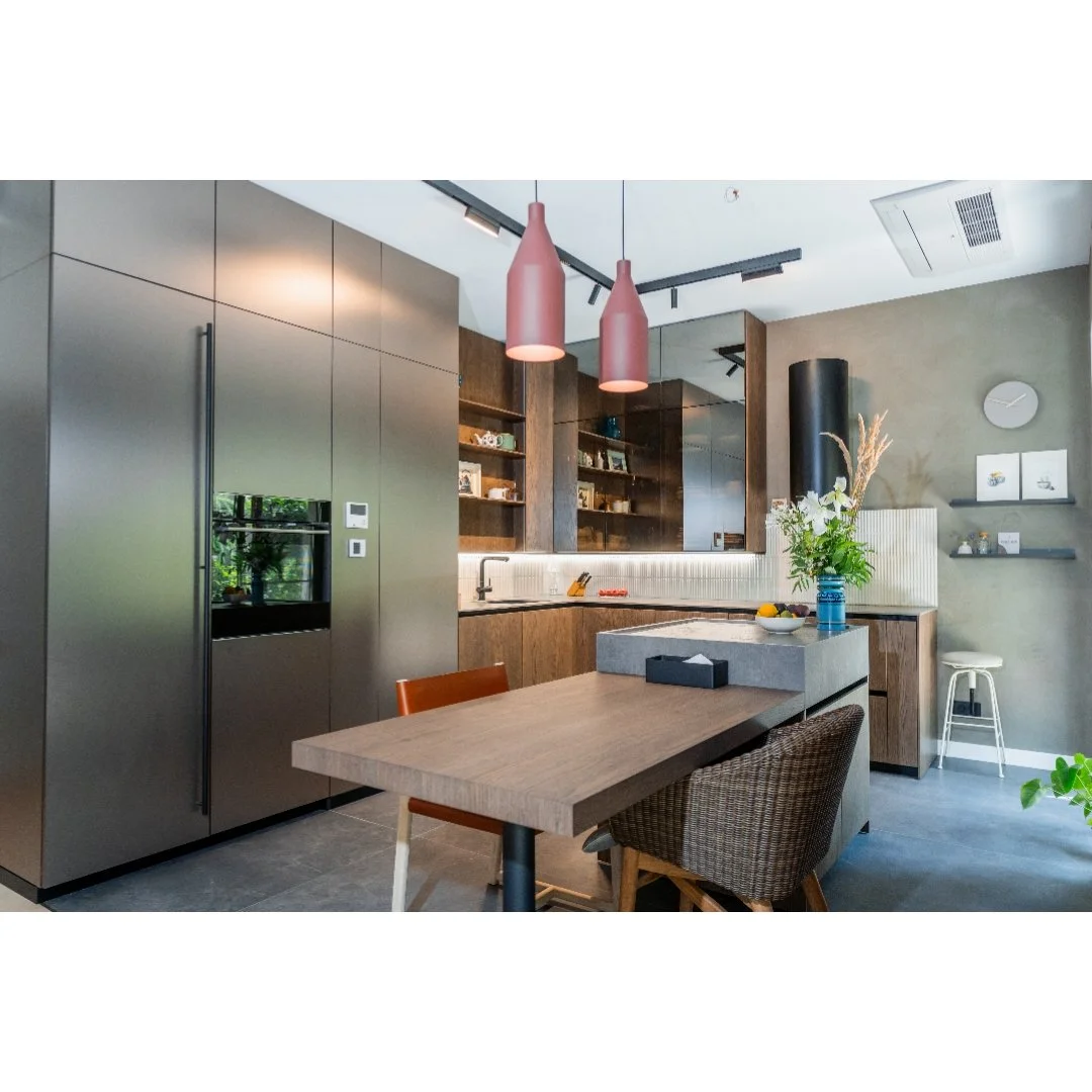 Modern kitchen with gray and wooden cabinetry, pink pendant lights, a wooden dining table, wicker and wooden chairs, a kitchen island with a plant, open shelves with decor, and a clock on the wall.