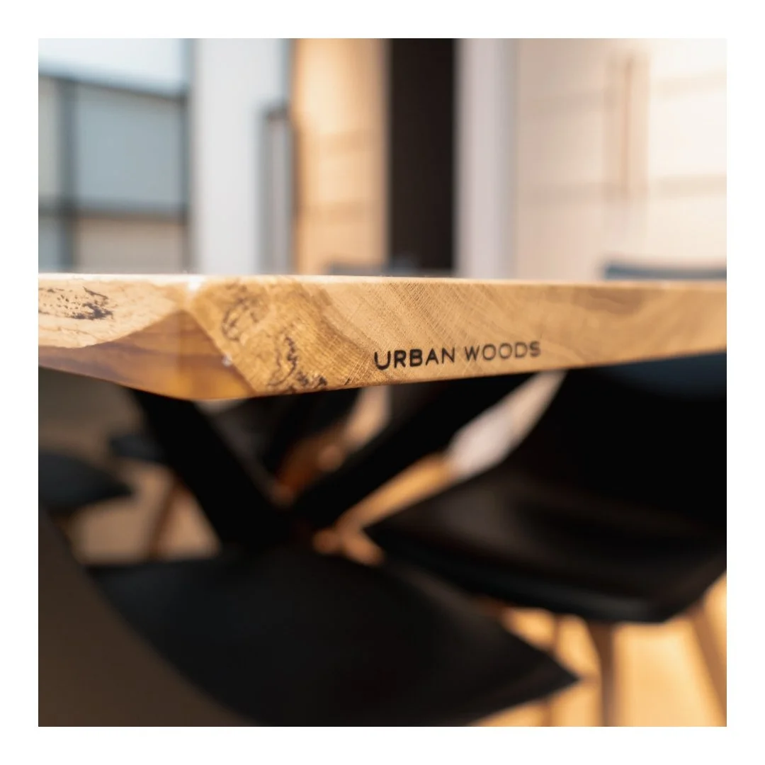 Close-up of a wooden table edge with the branding 'URBAN WOODS' engraved, with a blurred background.