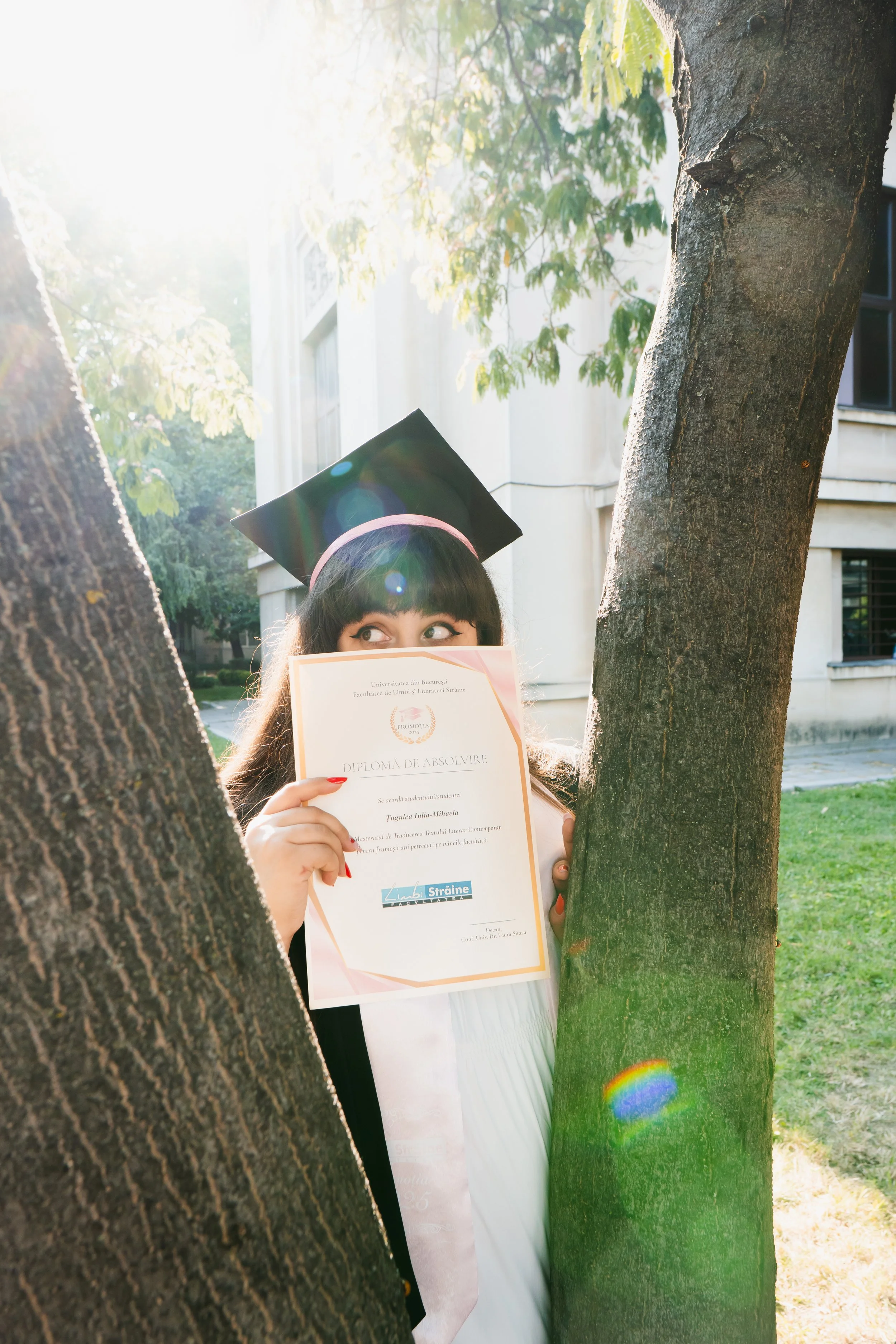 A young woman in a graduation cap holding a diploma, standing between two trees outdoors with sunlight filtering through the leaves.