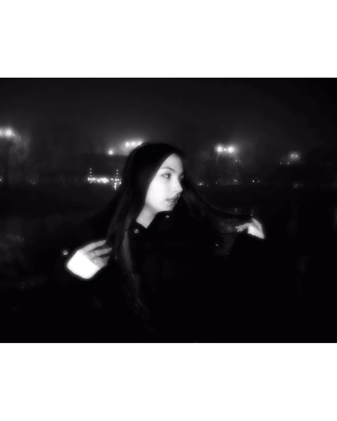 A woman with long dark hair in a dark coat looks to the side at night in a foggy city setting.