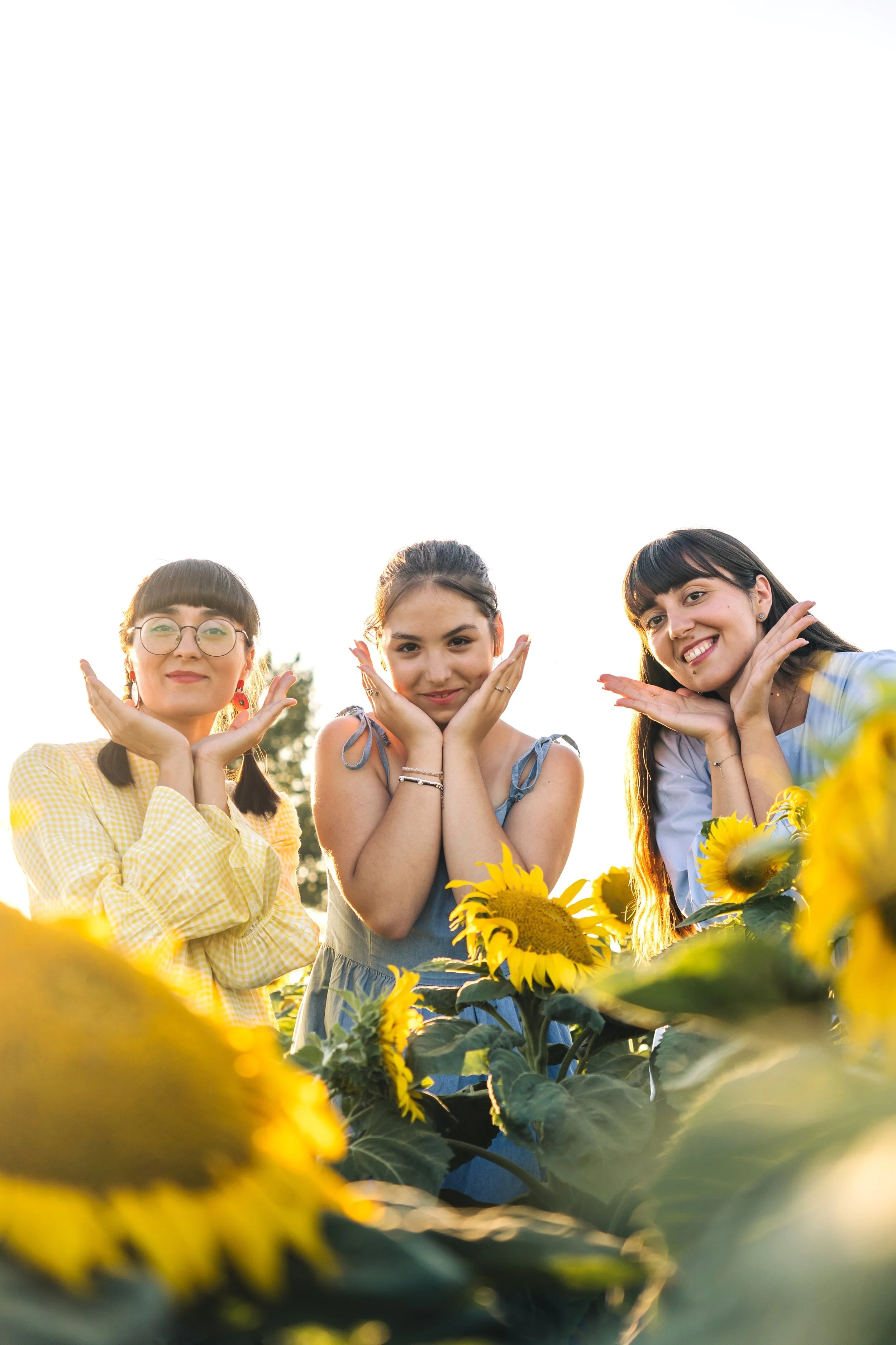 Three young women posing among sunflowers during sunset, making playful faces and gestures.