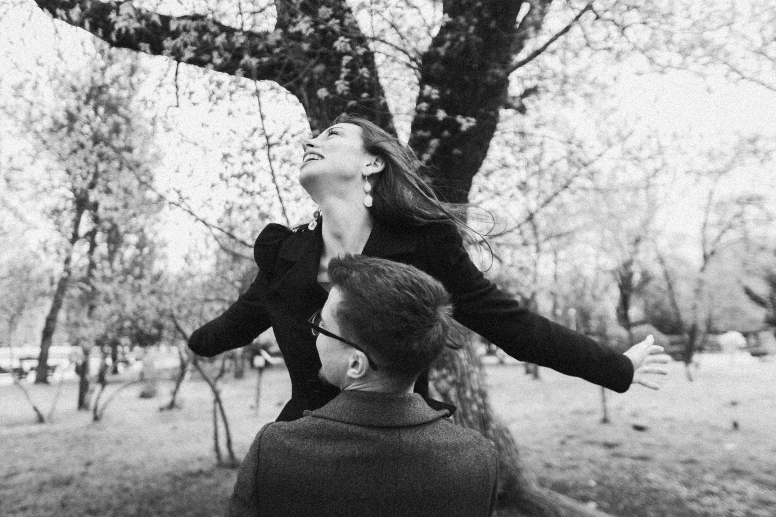 A woman with earrings smiling and looking up while being lifted by a man with glasses outside in a park with trees.