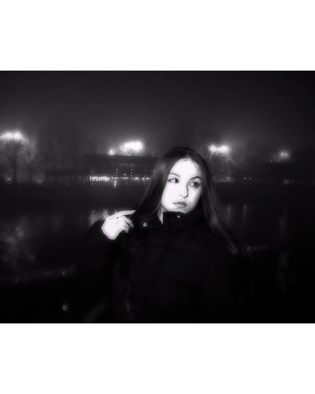 A black and white photo of a young woman outdoors at night, standing near a body of water with lights reflecting on it, with trees in the background.