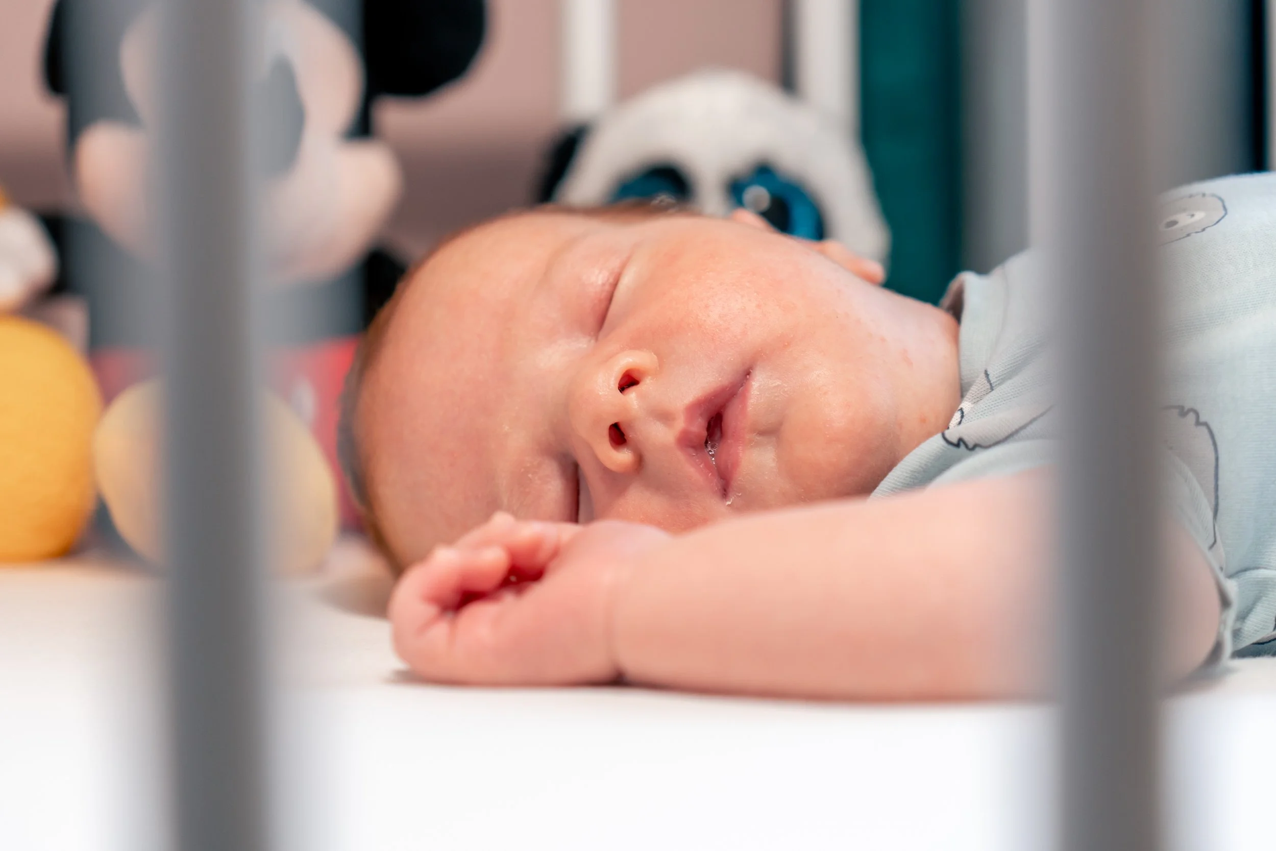 Close-up of a sleeping baby lying on a white surface, with toys nearby, surrounded by a metal railing.