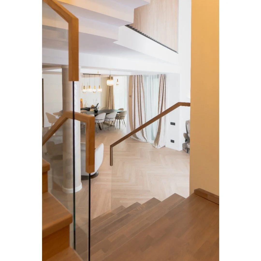 Interior view of a modern home with wooden staircase, dining area with hanging lights, large curtains, and a light-colored hardwood floor.