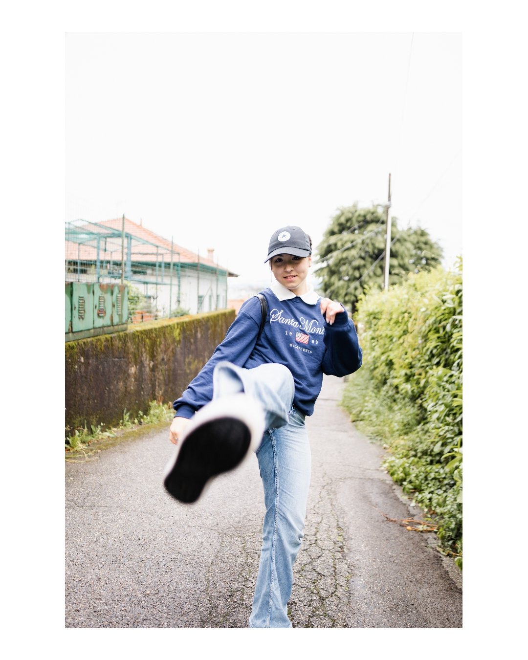A young woman kicking towards the camera on a narrow outdoor path, wearing a navy sweatshirt, light blue jeans, a black cap, and carrying a backpack, with greenery on both sides.