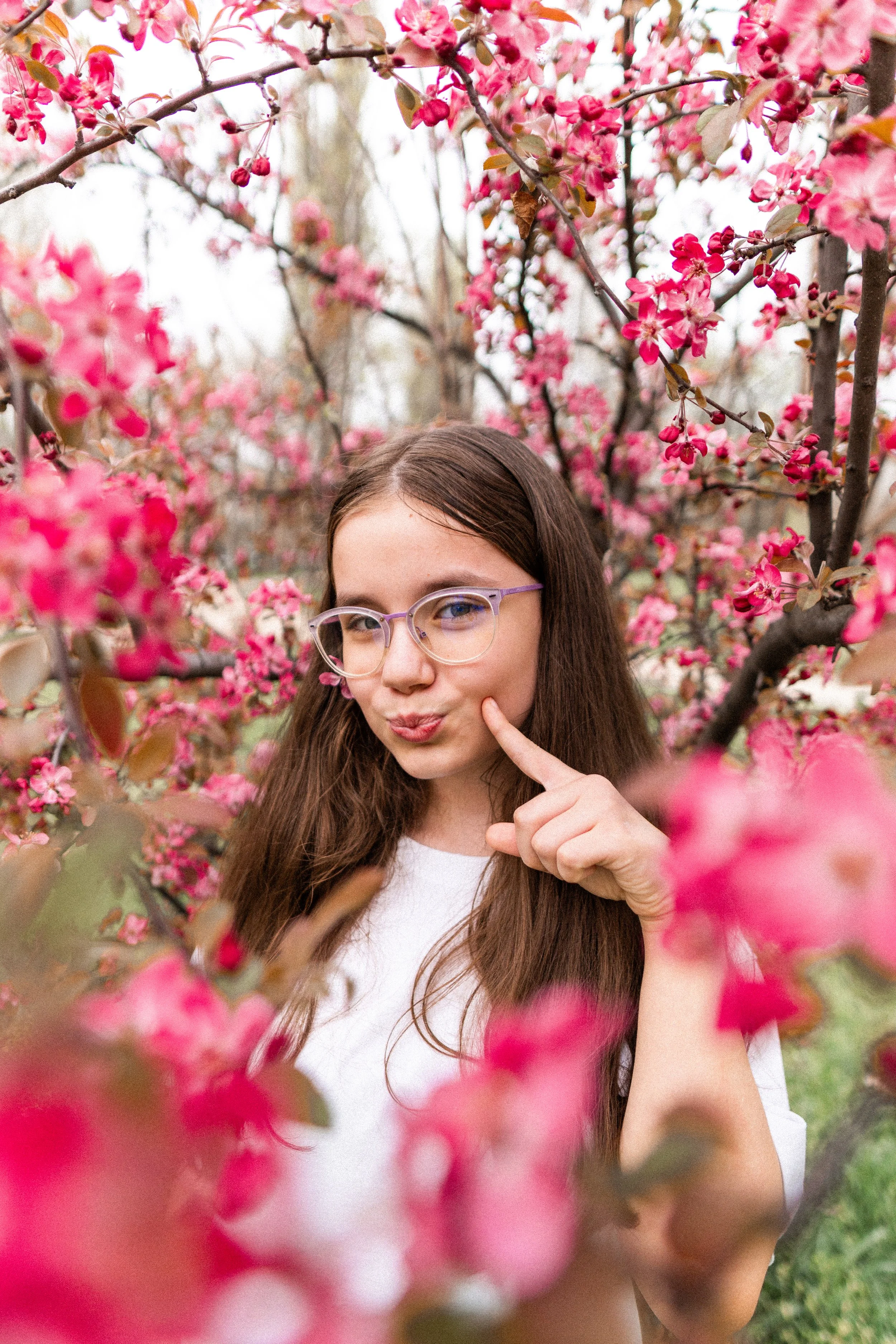 A woman with glasses posing playfully among blooming pink flowers on trees.