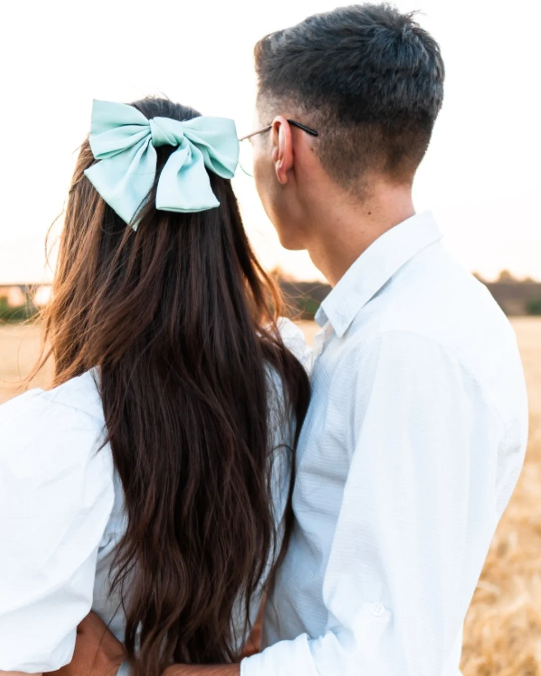 A couple standing closely together outdoors during sunset, with the woman having long, wavy brown hair with a large light blue bow and the man with short dark hair and glasses, both wearing white shirts.