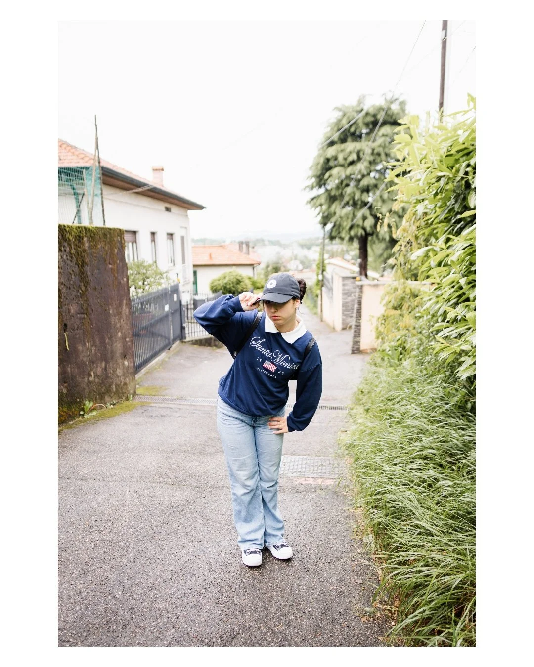 A young woman standing on a residential street, wearing a dark blue sweatshirt, light blue jeans, white sneakers, a dark cap, and a backpack, adjusting her cap.