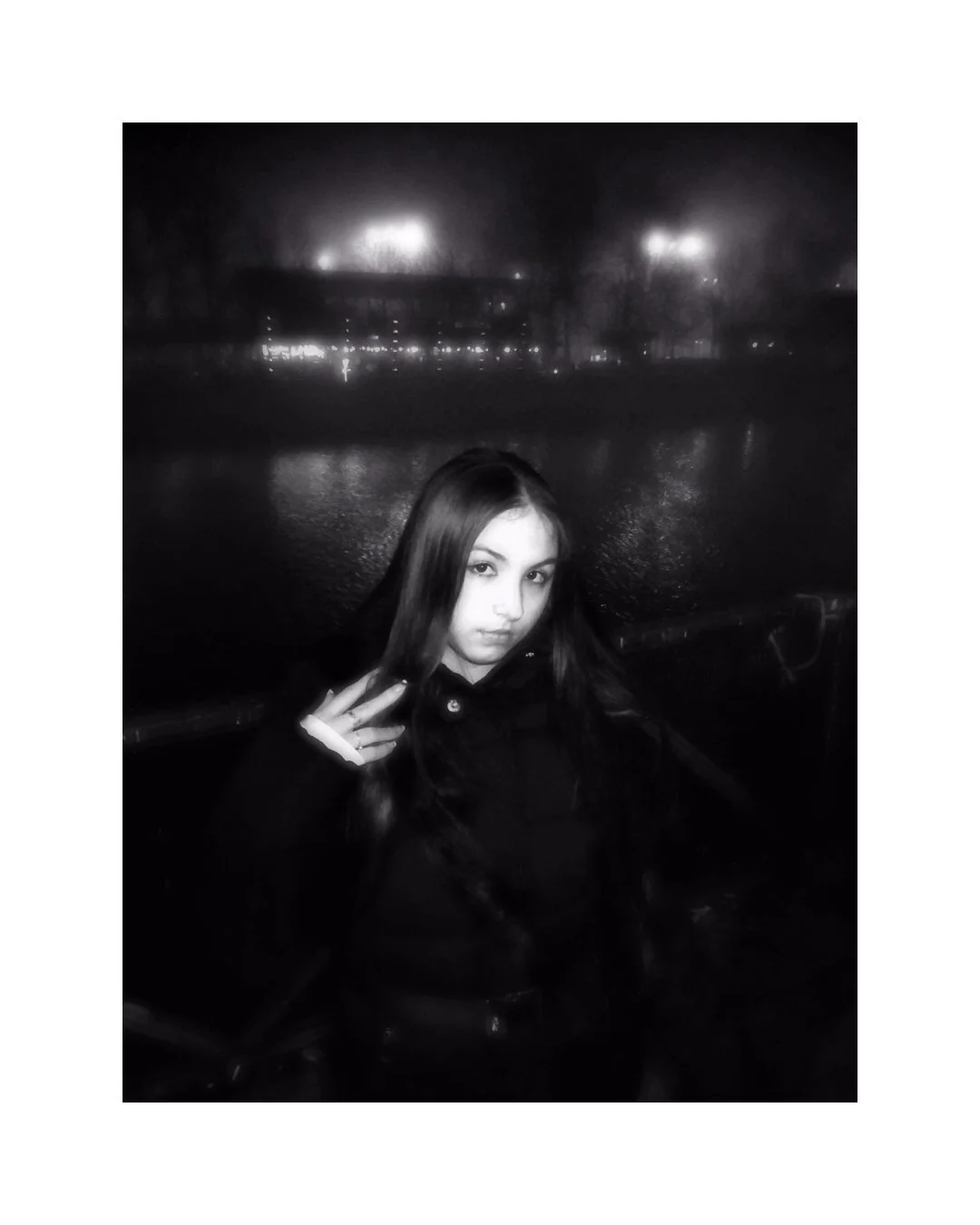 A young woman with long dark hair making a peace sign, posing outdoors at night with a waterfront and city lights in the background, in black and white.