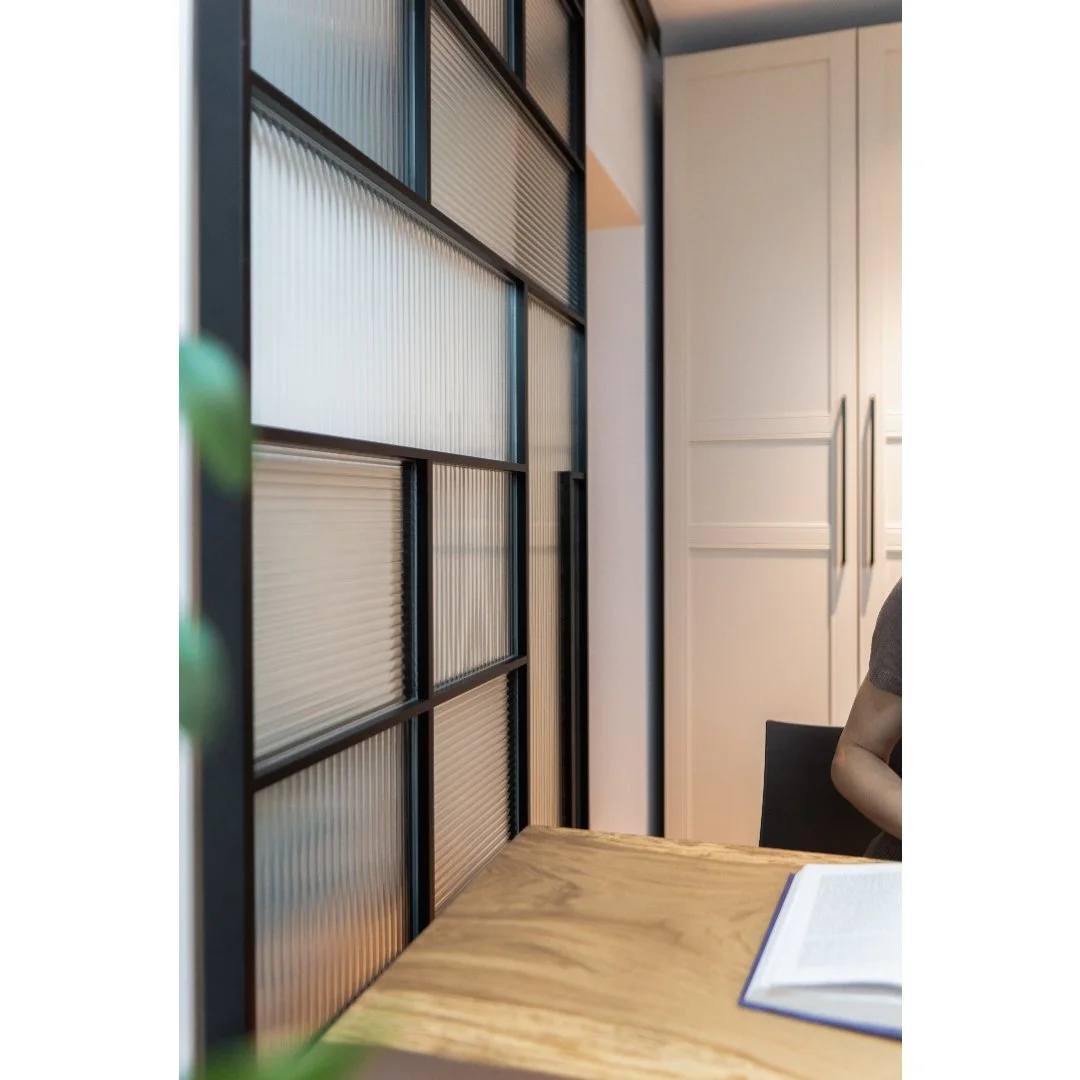 Partial view of an interior space showing a black metal and glass partition, a wooden table with an open book, and cabinets with vertical handles in the background.