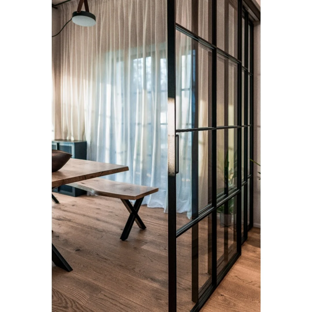 Interior of a modern room with wooden flooring, a wooden table with a bench, sheer white curtains with sunlight streaming in, and a black metal-framed glass partition wall.